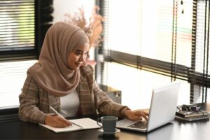 A woman leader sits at her desk writing in a notebook while working on a laptop, preparing a self-evaluation for her performance review.