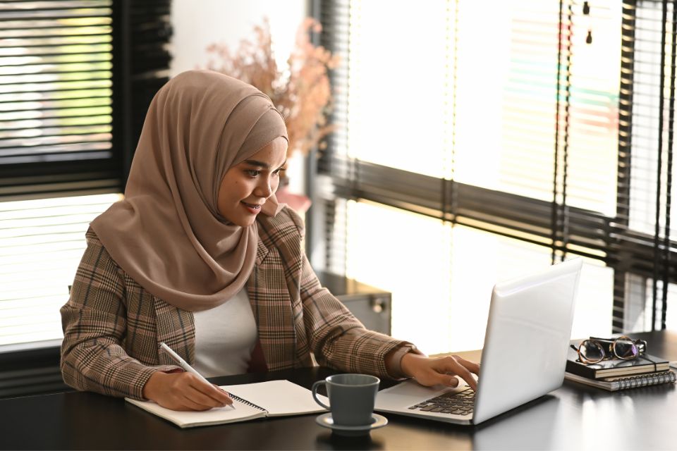 A woman leader sits at her desk writing in a notebook while working on a laptop, preparing a self-evaluation for her performance review.
